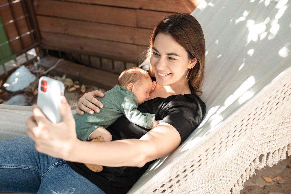 A heartwarming moment of a mother cuddling her baby on a hammock while capturing a selfie.
