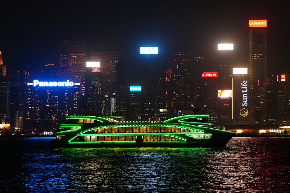 A neon-lit ferry cruises in front of the vibrant Hong Kong cityscape at night.