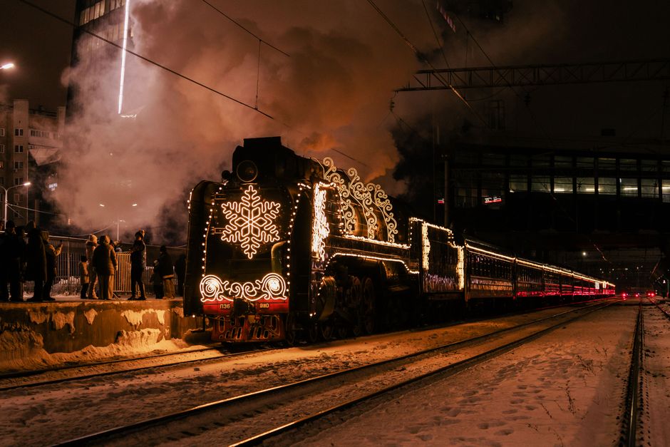 A festive steam train decorated with lights, emitting steam at a snow-covered station during nighttime.