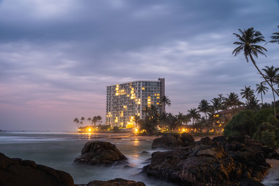 Stunning beachside hotel illuminated against dusk sky with palm trees and rocky shores.