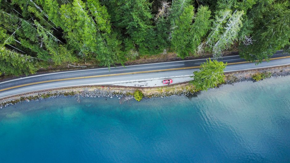 Aerial view of a car on a road by a tranquil lakeside surrounded by lush green forest trees.