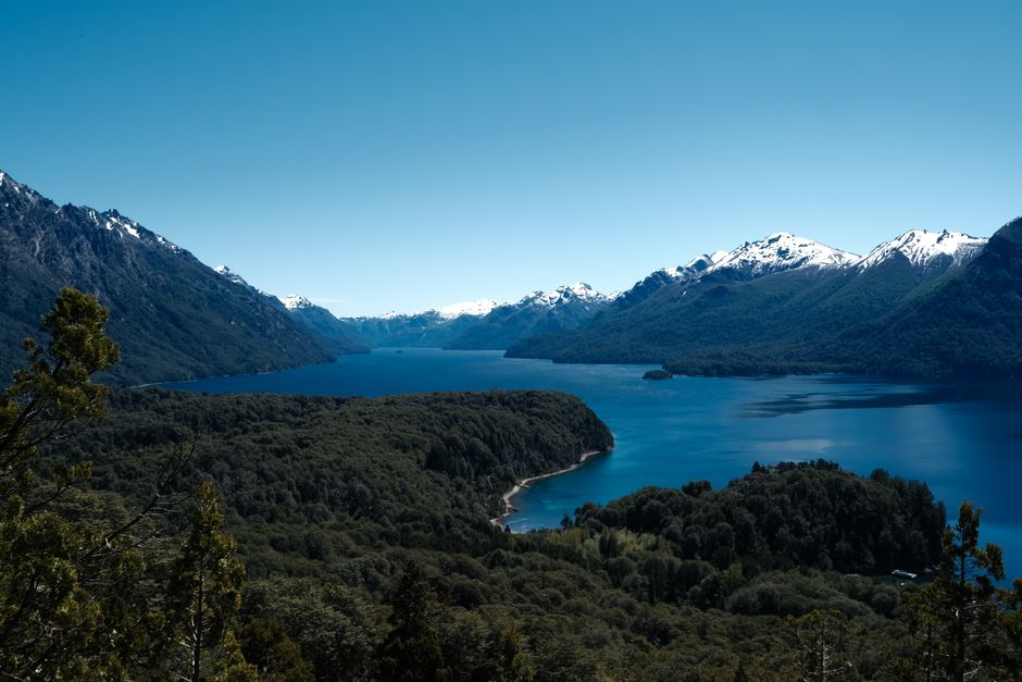 Lush forest and snowy mountains surround the clear blue waters of Nahuel Huapi Lake in Patagonia, Argentina.