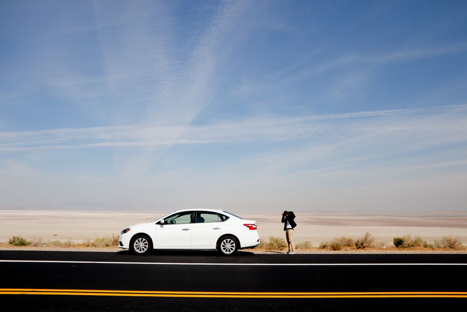 A white car parked on a desert road with a photographer capturing the vast landscape under a clear sky.