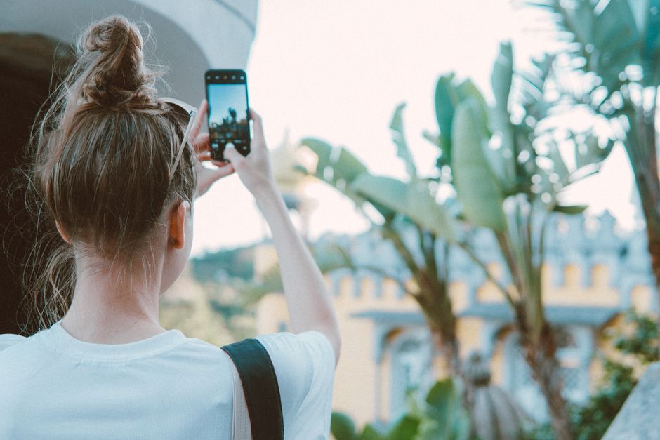 A woman takes a smartphone photo of a palace surrounded by banana trees in Sintra, Portugal.