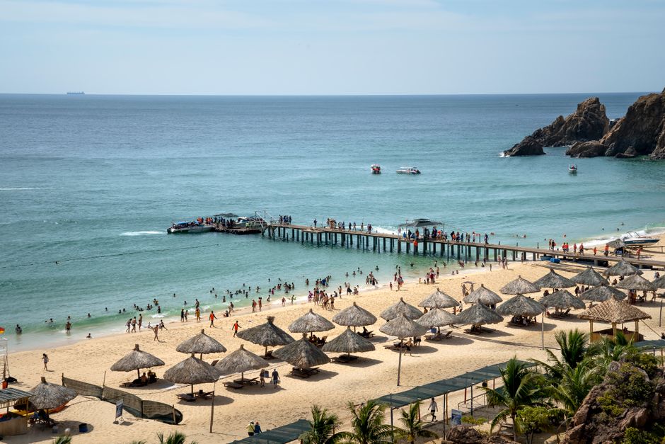 People chilling on spacious sandy beach with straw umbrellas and narrow wharf near turquoise rippling sea on sunny day